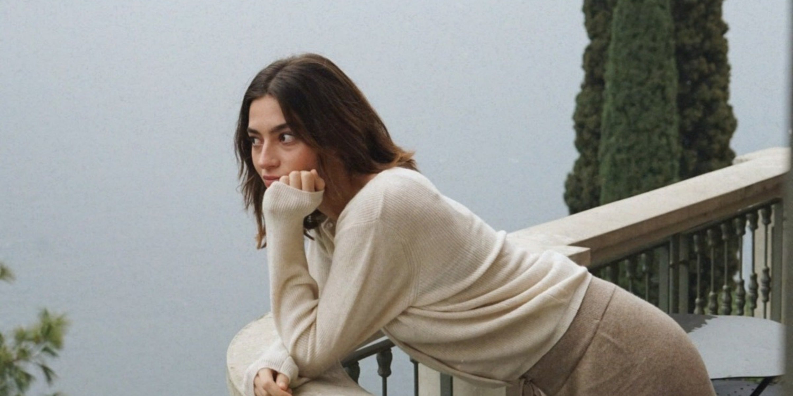 A woman in a cream sweater leaning on a balcony railing with trees and pale water in the background.