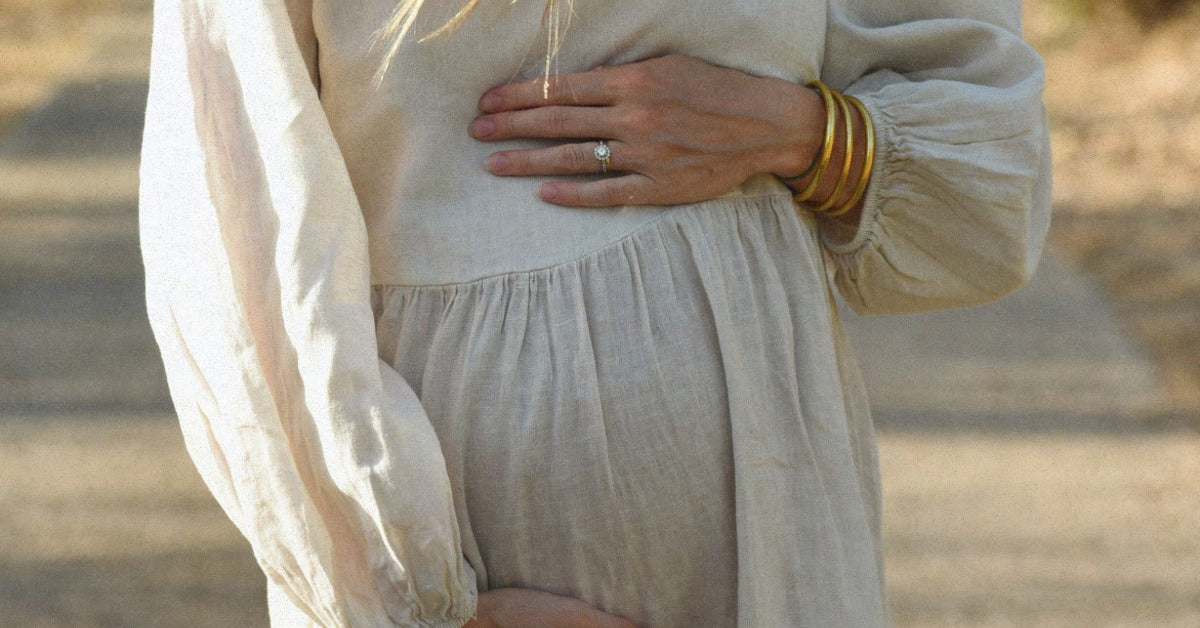 A woman in a cream color, long-sleeve dress holding her rounded abdomen while standing outdoors among trees.