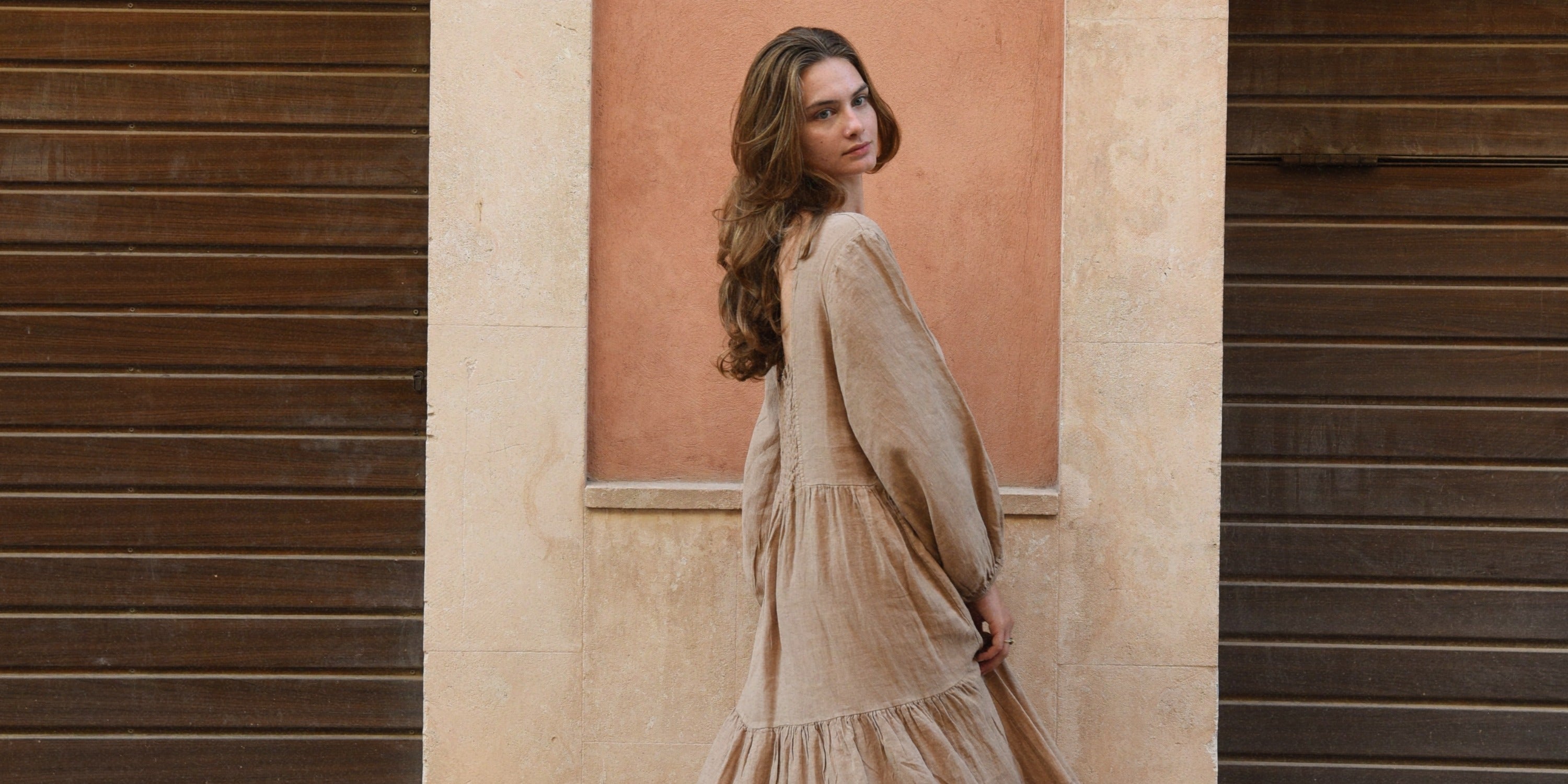 A woman in a beige kaftan walking beside a peach wall between two dark wooden doors and looking over her shoulder.
