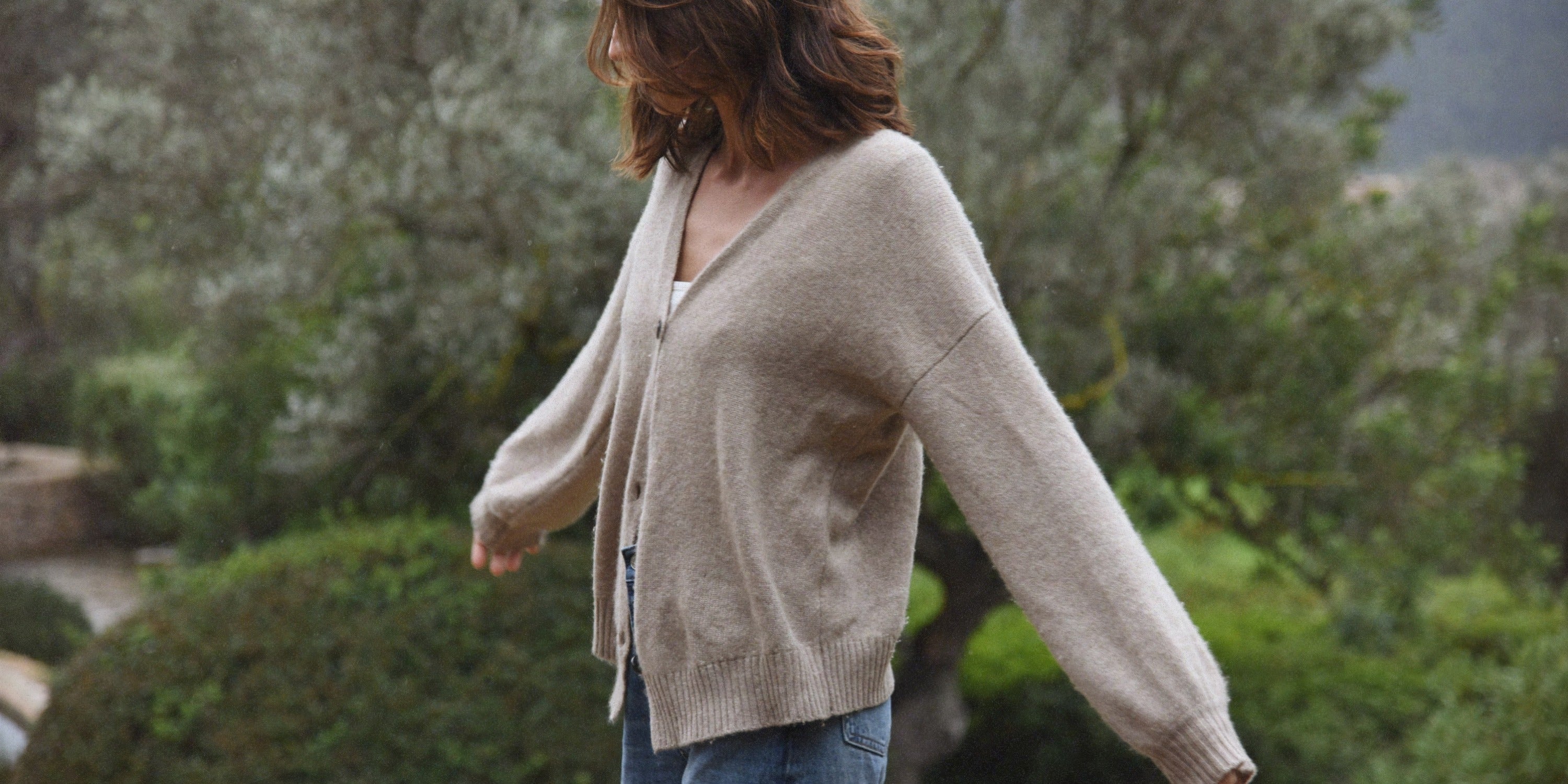 A woman in a beige knit cardigan and jeans stands outdoors with her arms relaxed, surrounded by soft green trees.