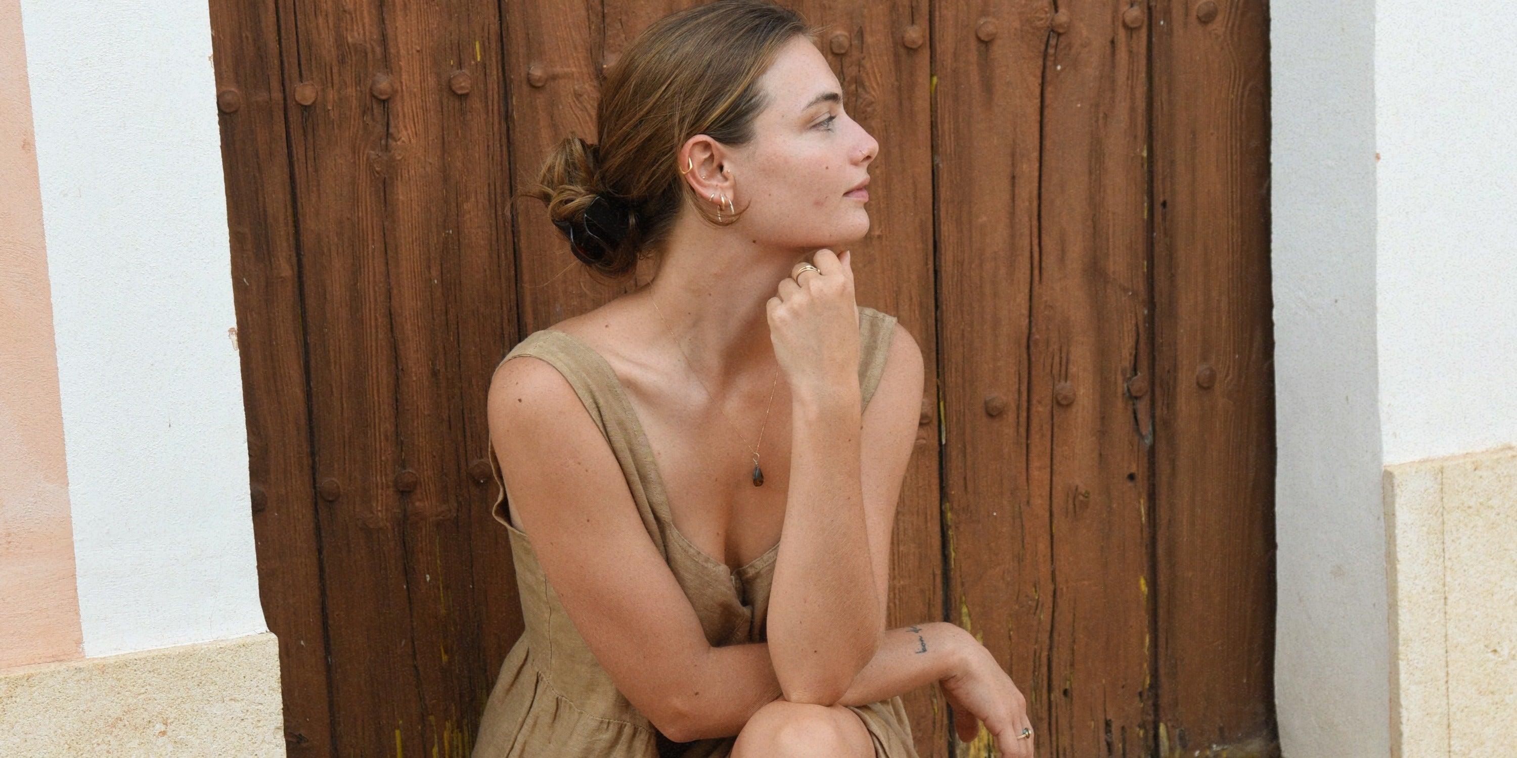 A woman sitting in front of a large wooden door with her hair tied back and one hand resting under her chin.