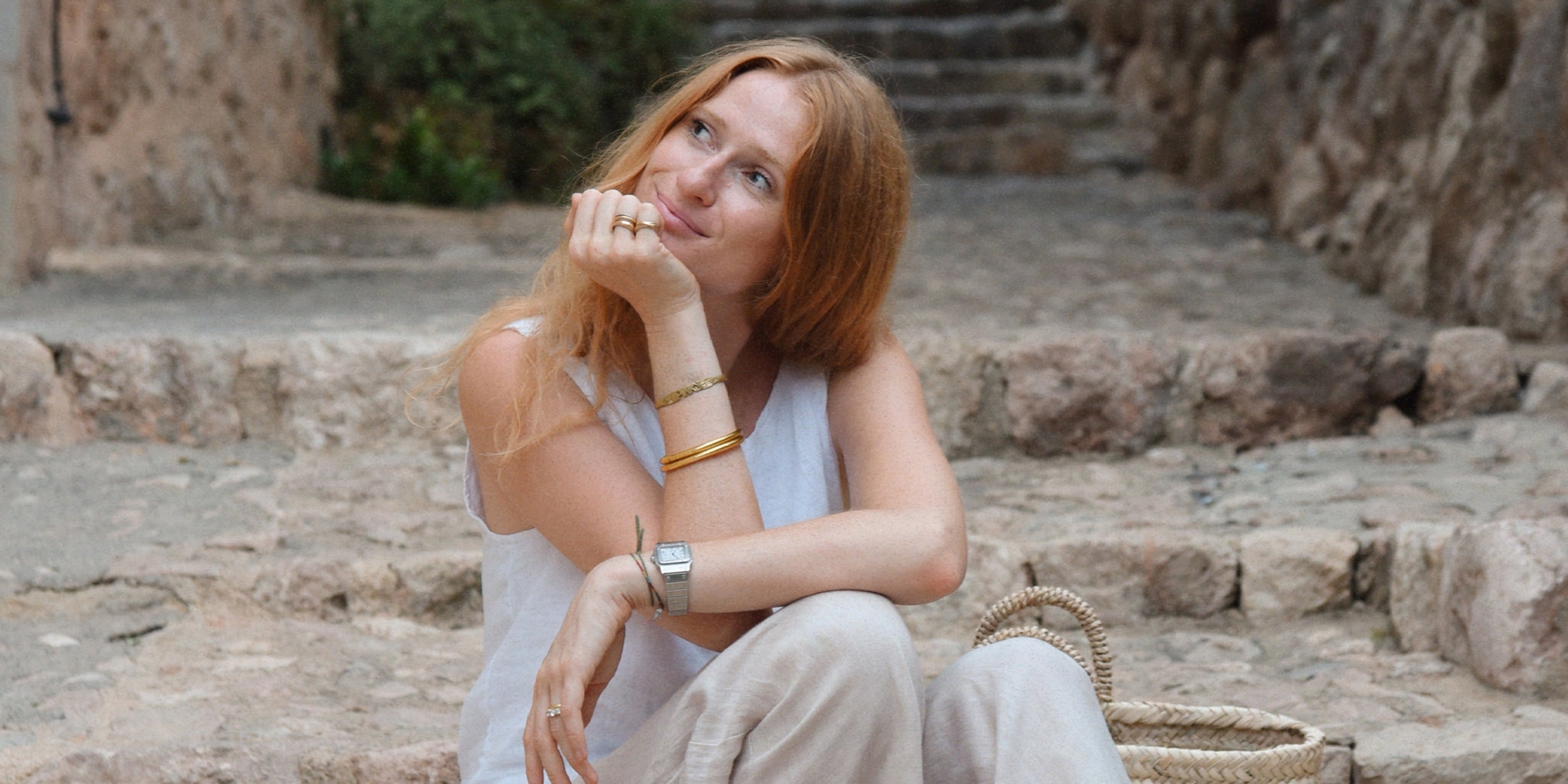 A woman with red hair sitting on stone steps in a narrow alley, wearing light linen clothes and tan sandals.