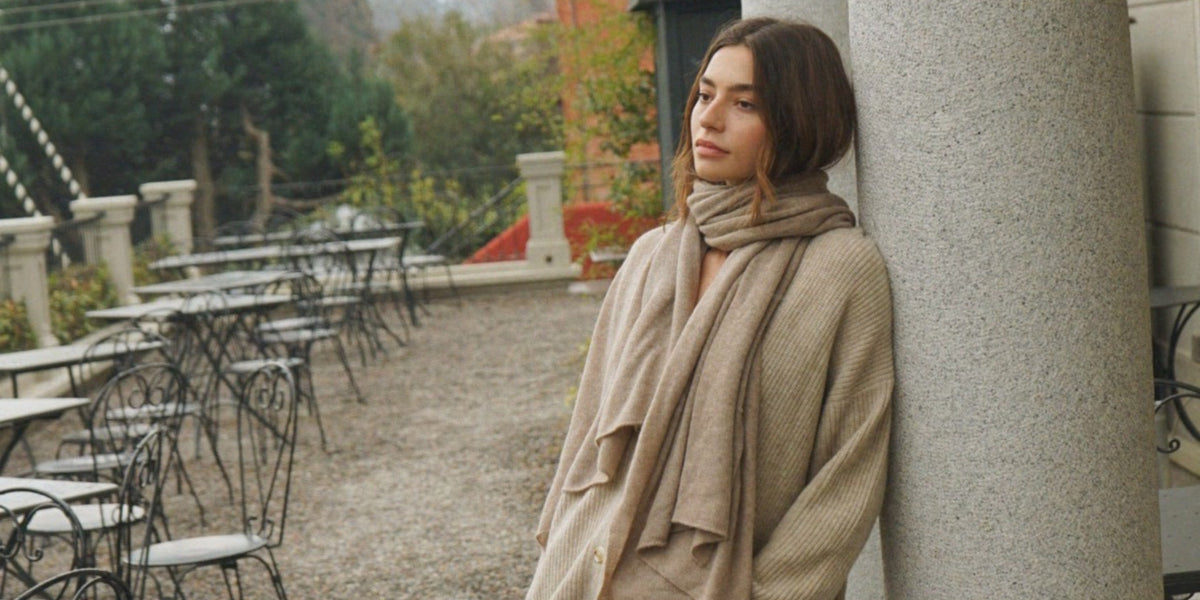 A woman wearing a beige scarf and knitwear leans against a stone column beside empty outdoor tables and chairs.