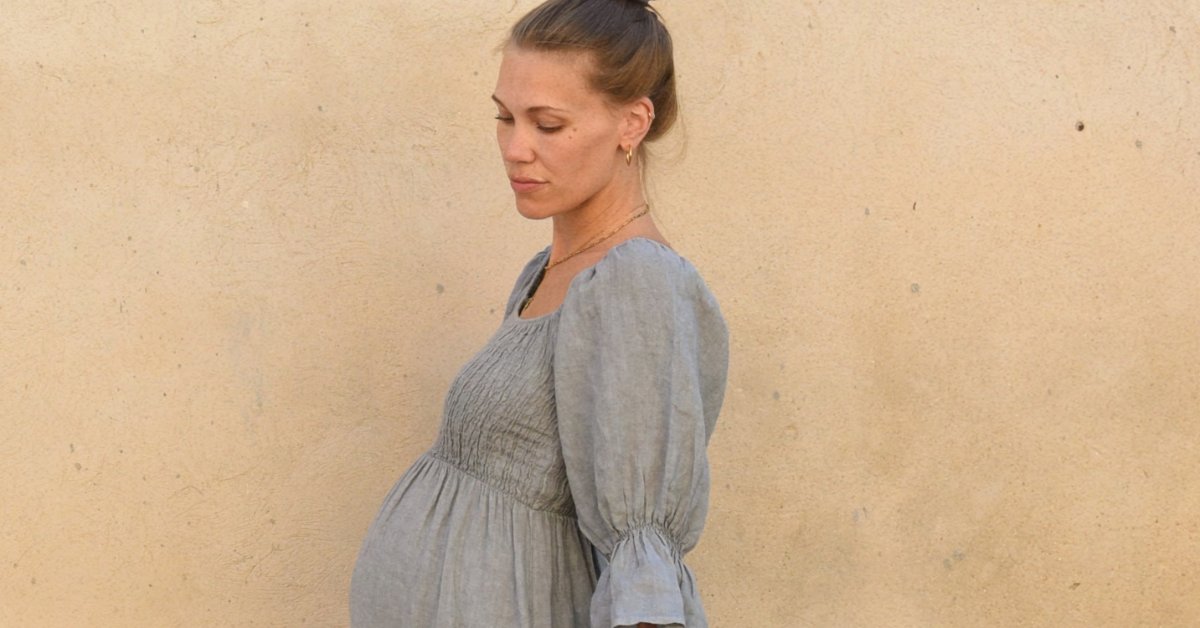 A pregnant woman in a blue dress standing sideways against a beige textured wall with her hair tied back.