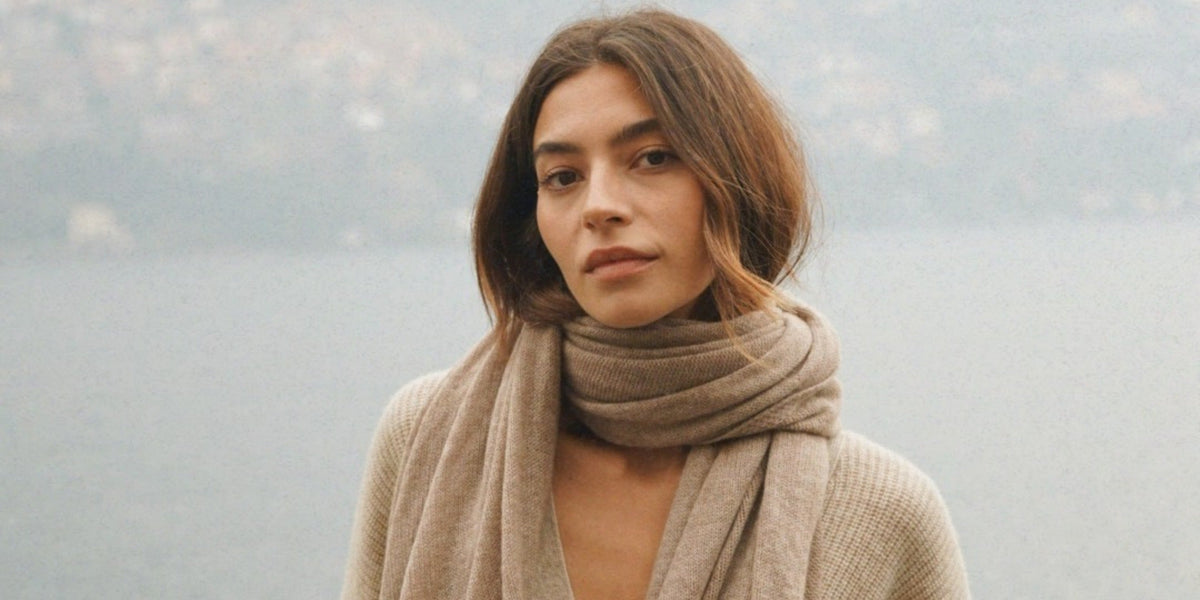 A woman wearing a thick beige scarf over a matching knit sweater standing outdoors against a misty waterfront.