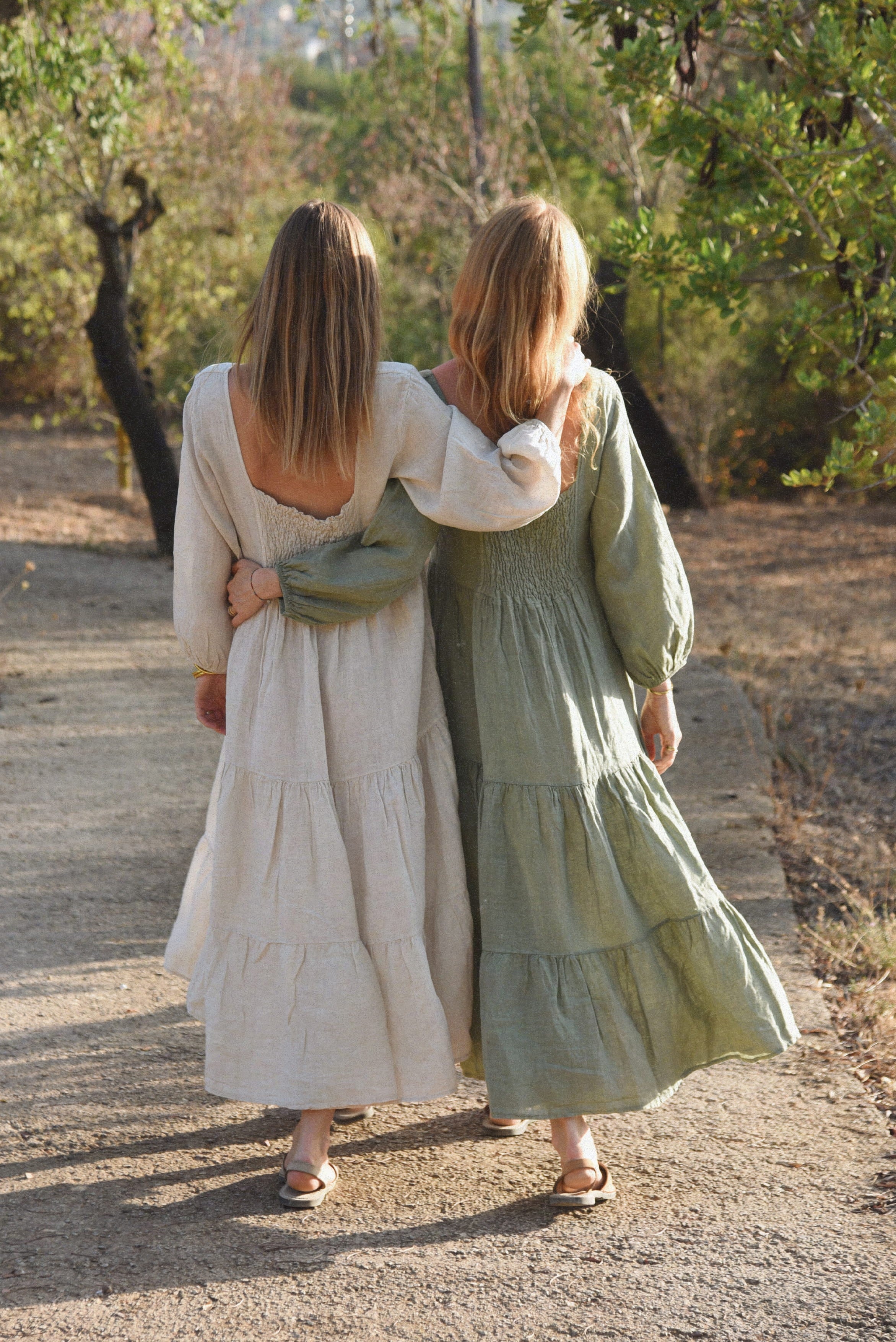 A rear view of two women in long dresses walking arm in arm down a sunlit dirt path bordered by trees.