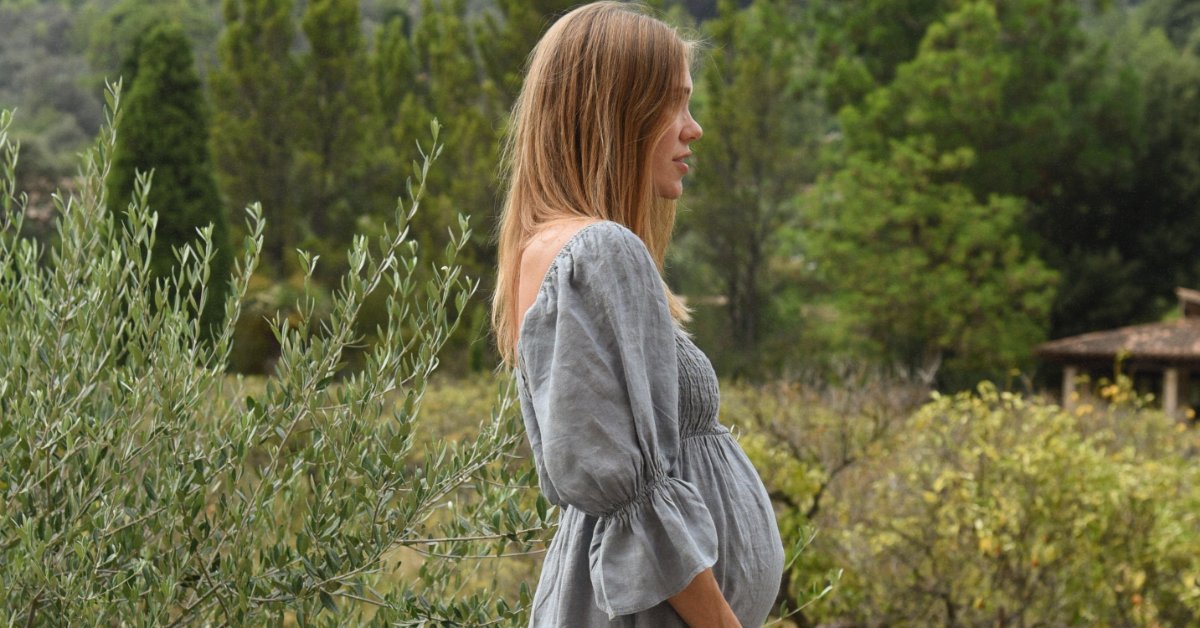 A pregnant woman in a gray dress standing barefoot beside shrubs with green hills and trees behind her.