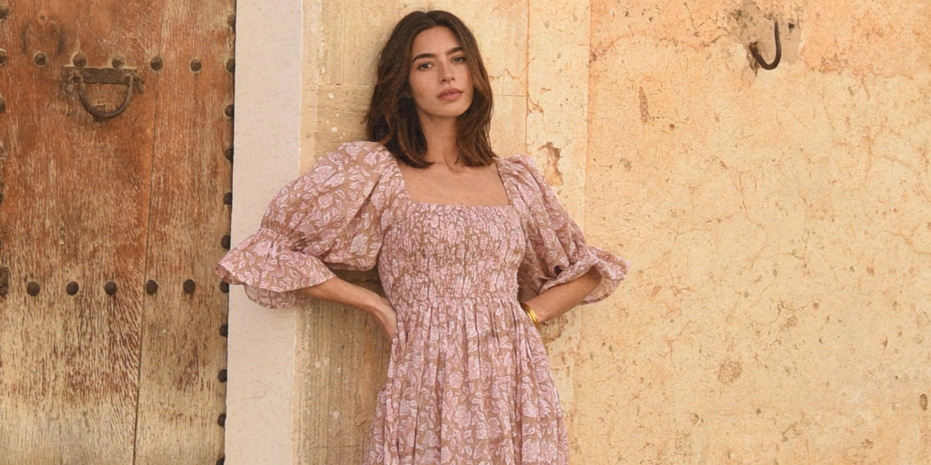 A woman in a pink floral dress leaning against a textured wall beside a wooden door with metal studs.