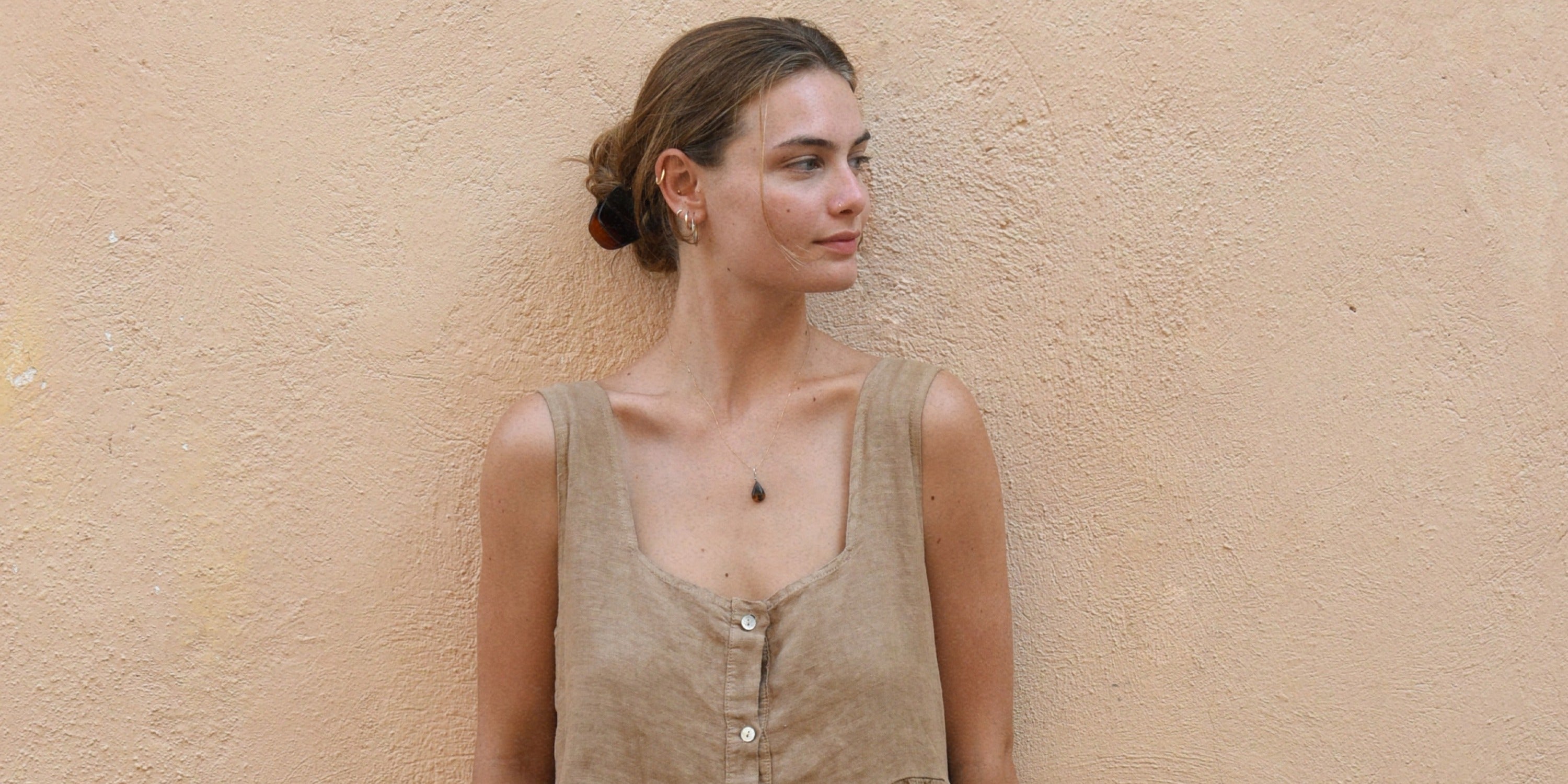 A woman in a tan sleeveless top standing against a textured beige wall, looking to the side with her hair tied back.