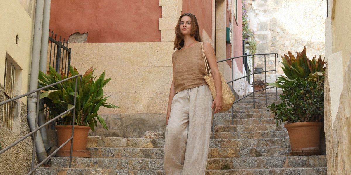 A woman wearing a beige sleeveless top and light pants, standing on stone steps beside potted green plants.