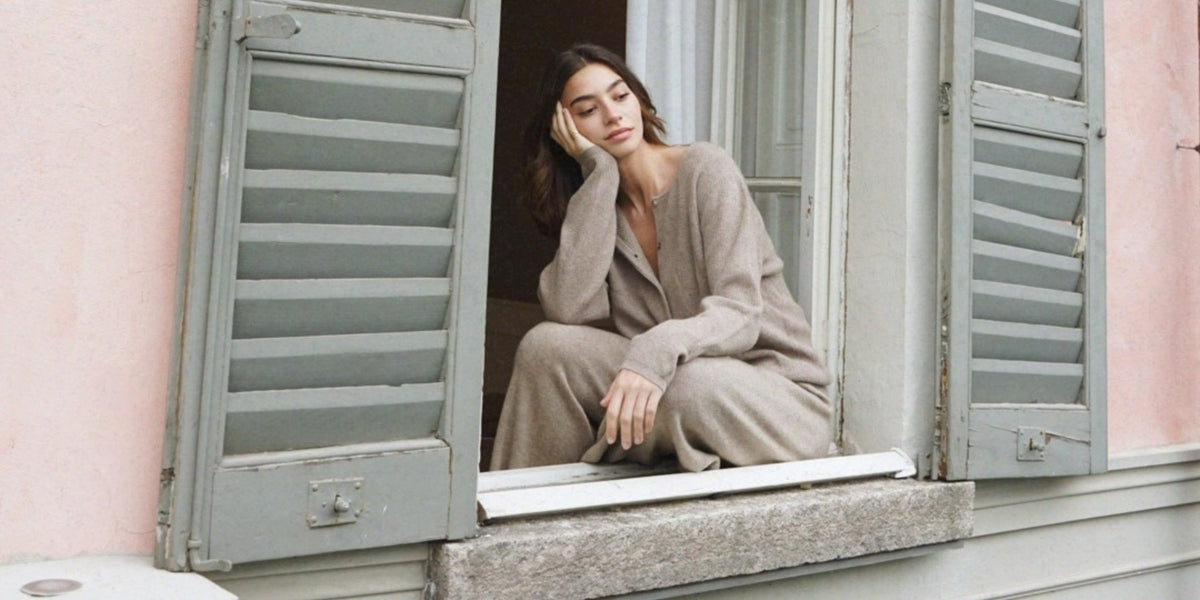 A woman in beige clothing sitting in an open window between pale green shutters and resting her head on one hand.