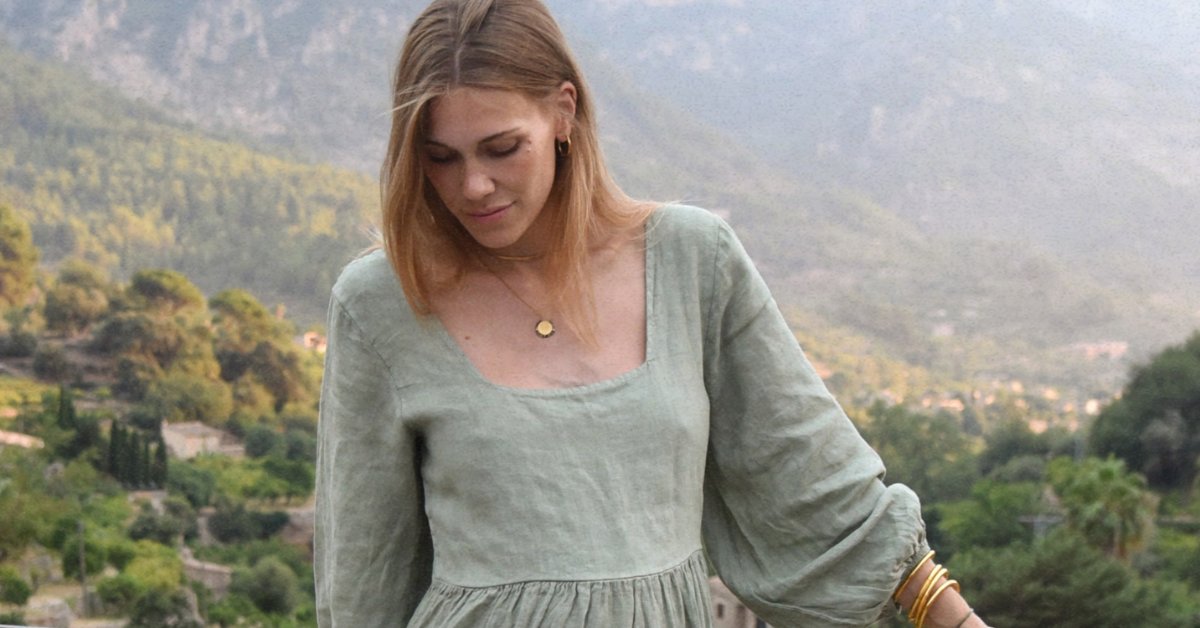 A woman in a pale green dress looking down with trees, hills, and distant mountains in the background.