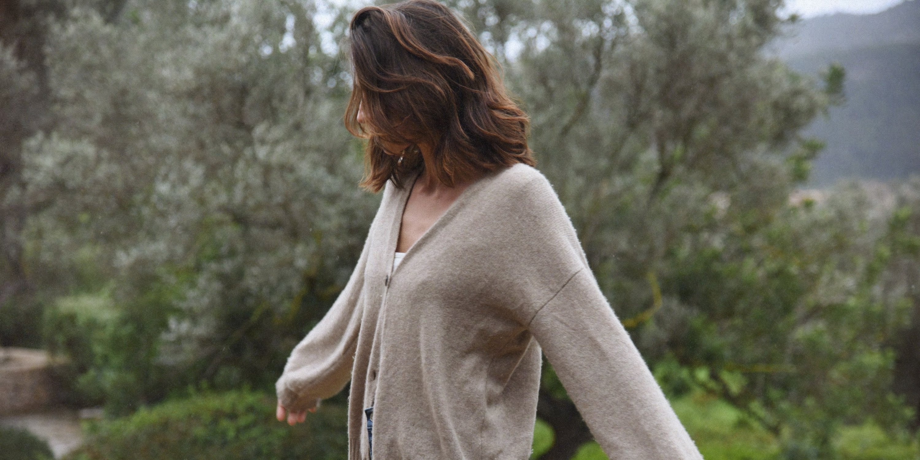 A woman walking outdoors beside green plants and trees while wearing a light cardigan over a white top.